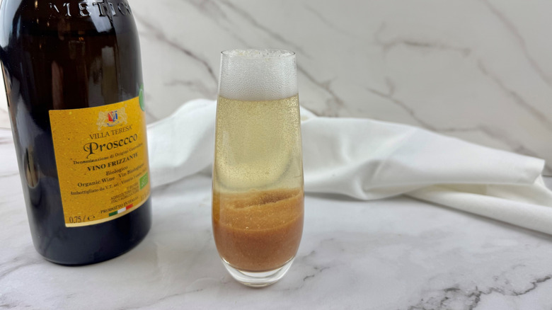 orange-colored cocktail in a drinking glass next to a crumpled napkin and a bottle of sparkling wine