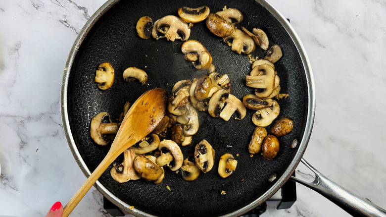 hand stirring mushrooms and minced garlic in frying pan with wooden cooking utensil