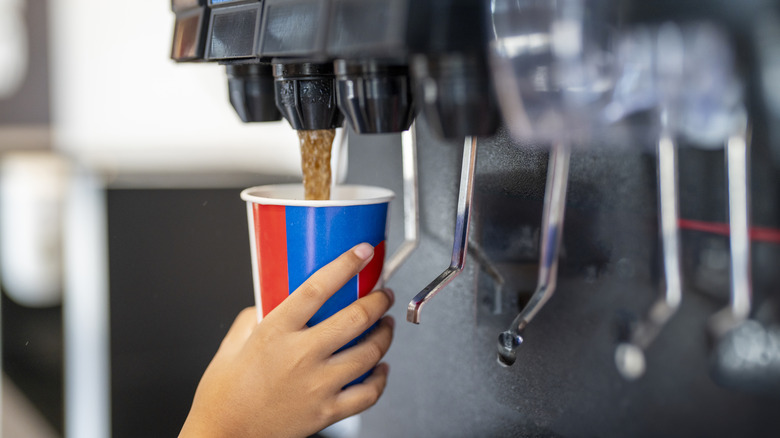 A hand fills a soda cup from a soda fountain