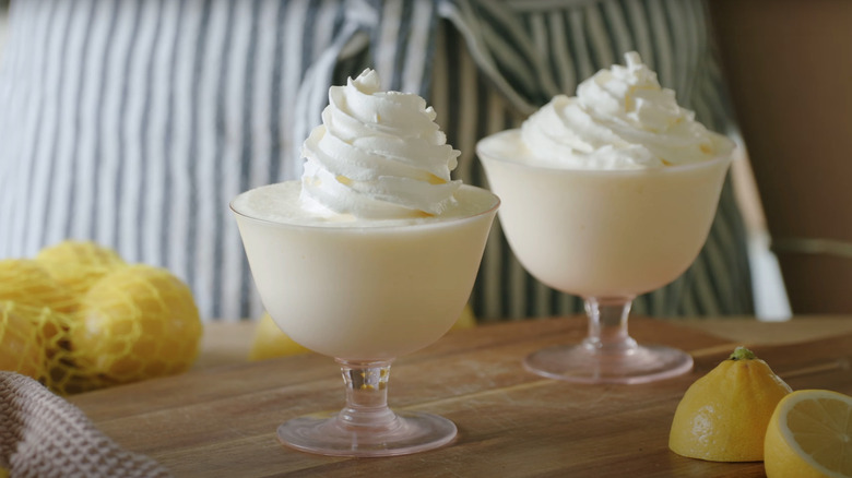 Two glasses of whipped lemonade topped with whipped cream sit on a counter by a bag of lemons