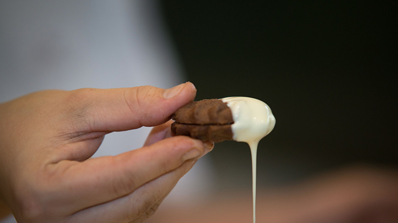 A chocolate cookie being dipped in a thin white chocolate coating