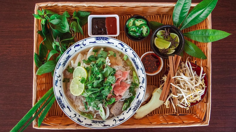 Pho in a blue and white bowl with sauces, sides, and greens placed in a woven basket