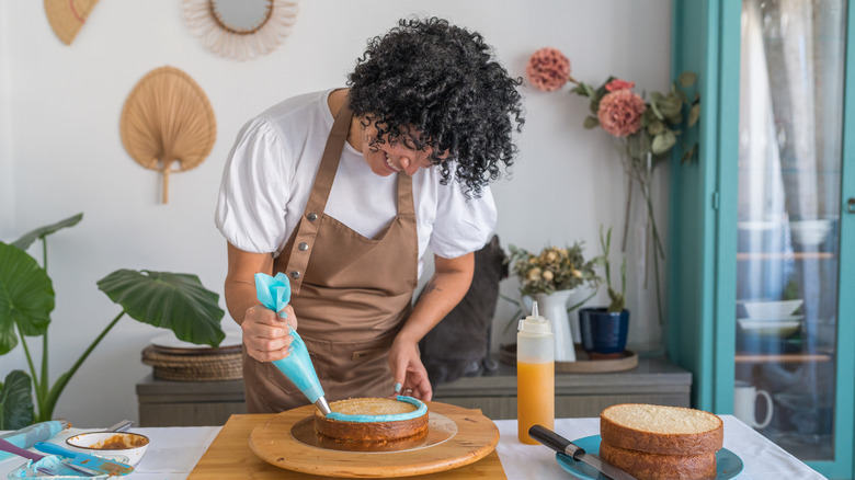 A baker in brown apron decorates a cake, squeezes out blue cream on a side of a cake